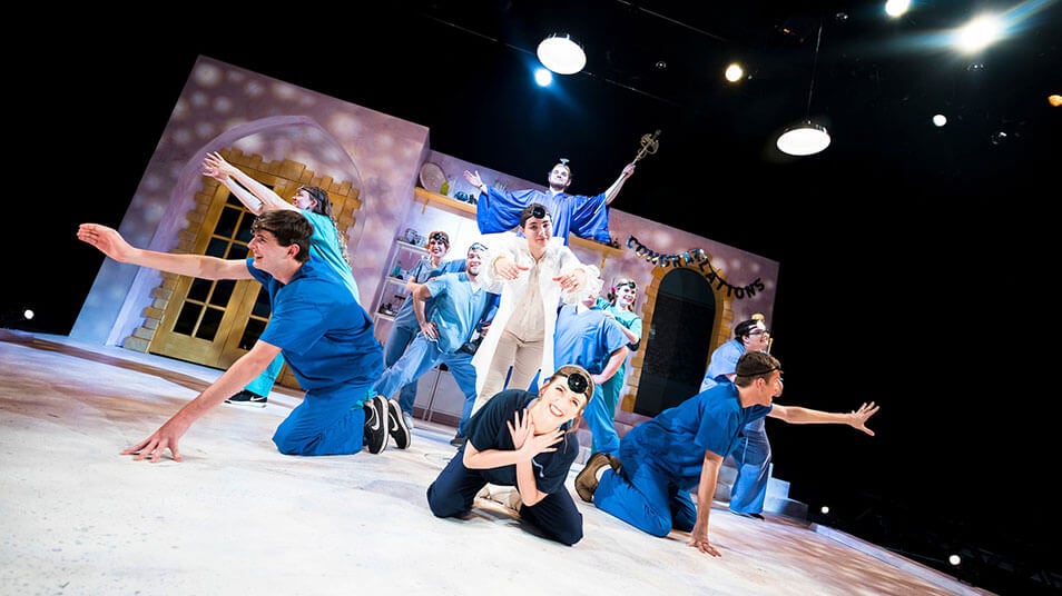 A group of students act out a scene in which one character dressed in all white is surrounded by blue costumed actors