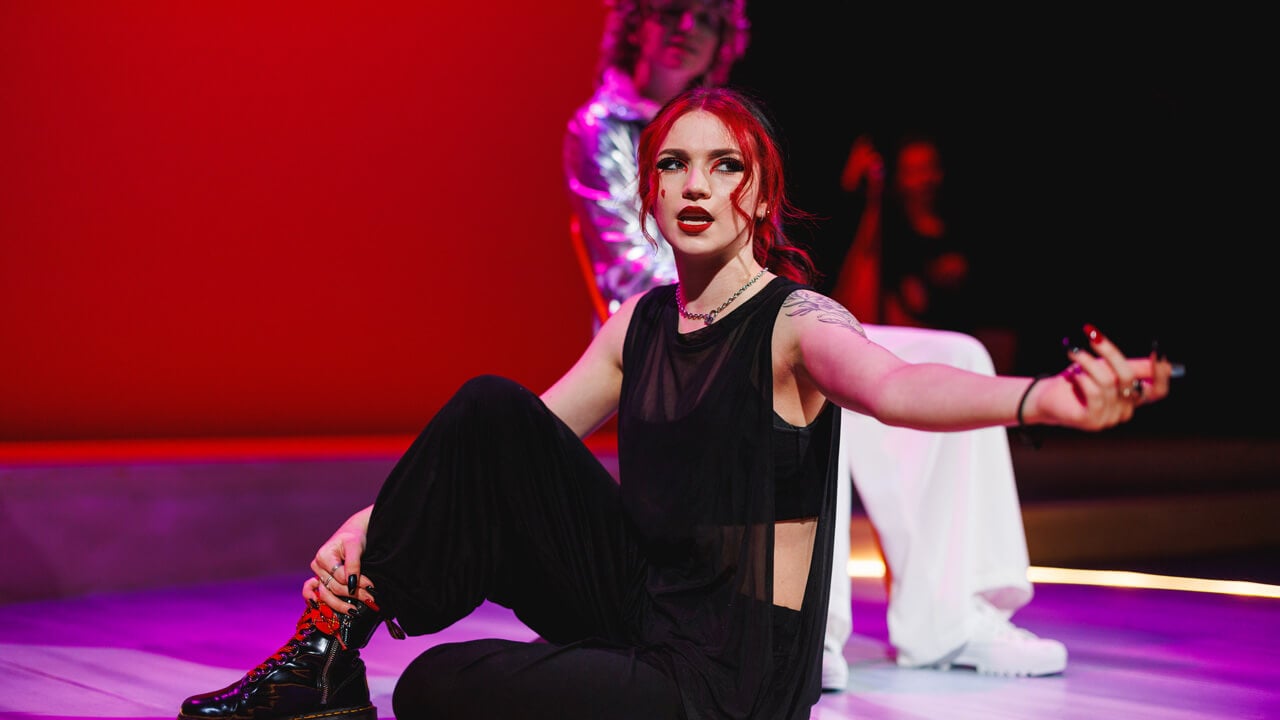 A female student actor on stage with red background lighting for the productions, Thebes