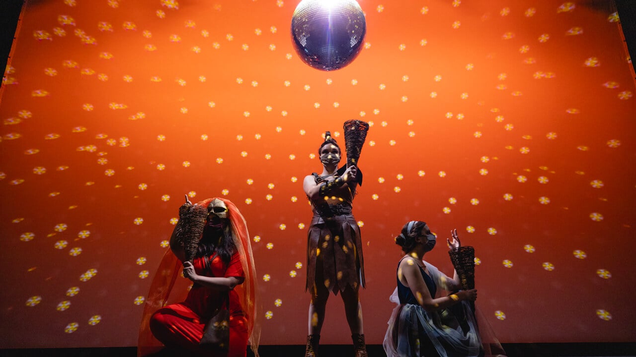 Three female student actors on stage for the production with orange lighting background, An Iliad