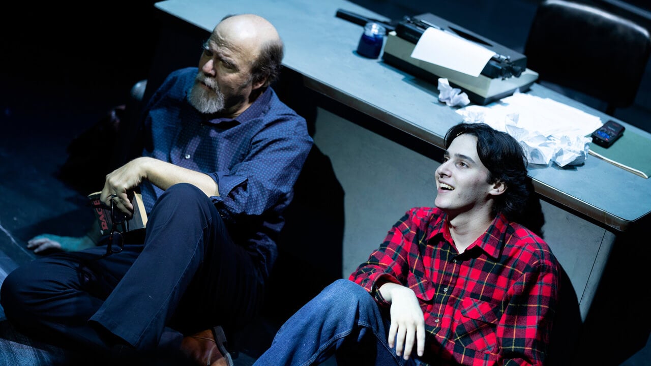2 male student actors on stage sitting on the ground leaning on a desk for the production, Rage
