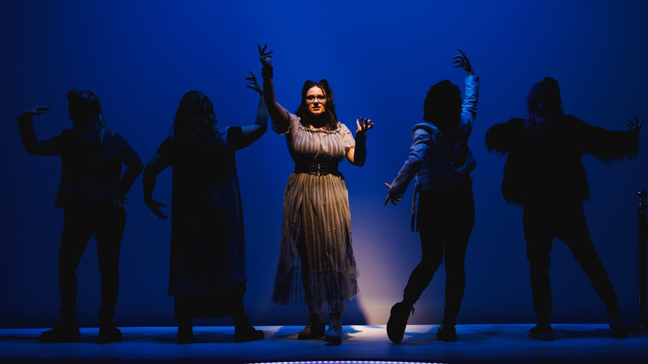 A group of student actors on stage singing with navy background lighting for the production, Thebes