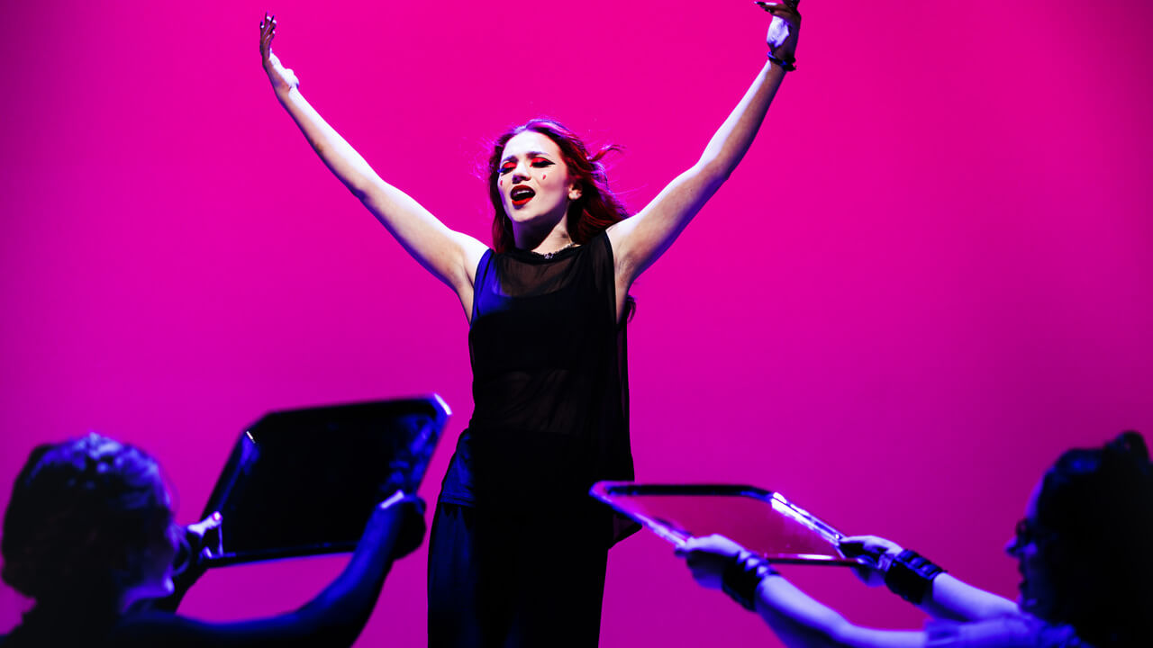 A student actor on stage singing with pink background lighting for the production, Thebes
