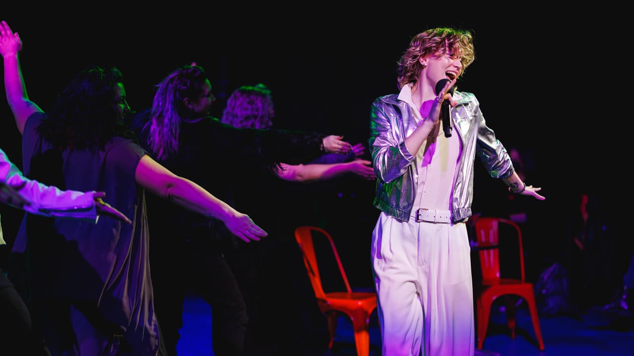 A student actor on stage singing with dark background lighting for the production, Thebes