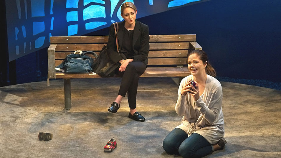 Two student actors in a scene where one sits on a park bench while the other kneels on the ground
